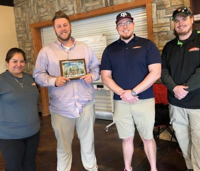 Three males and one female standing in office area with recently awarded Chamber of Commerce Member plaque.